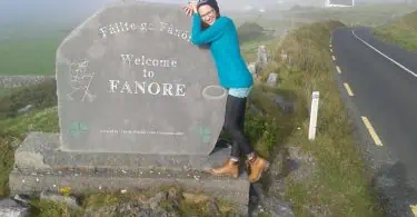 Ortseingangsschild von Fanore im Westen Irlands mit einer nebligen Landschaft im Hintergrund und Autorin Jessica Jirschik, die sich lächelnd an den Stein lehnt