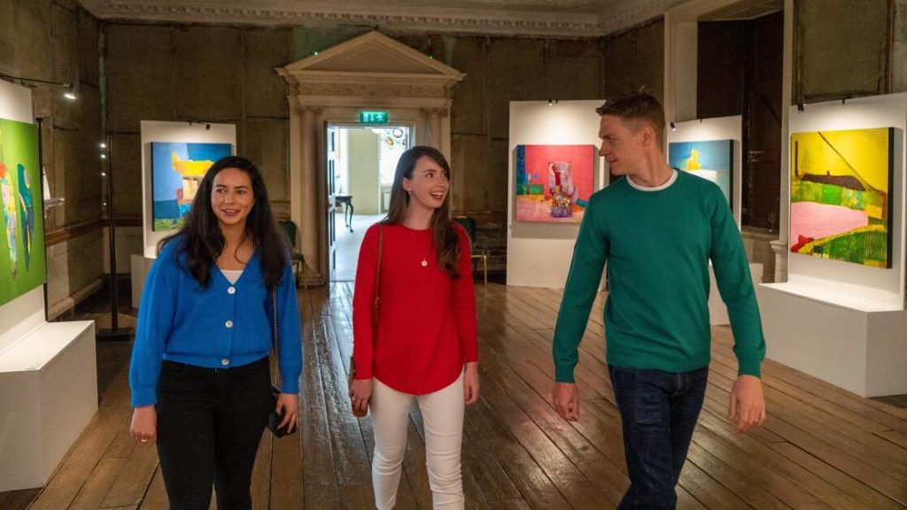 Drei junge Menschen schlendern lachend durch die Galerie des Rathfarnham castle. Im Hintergrund sind bunte Kunstwerke zu sehen